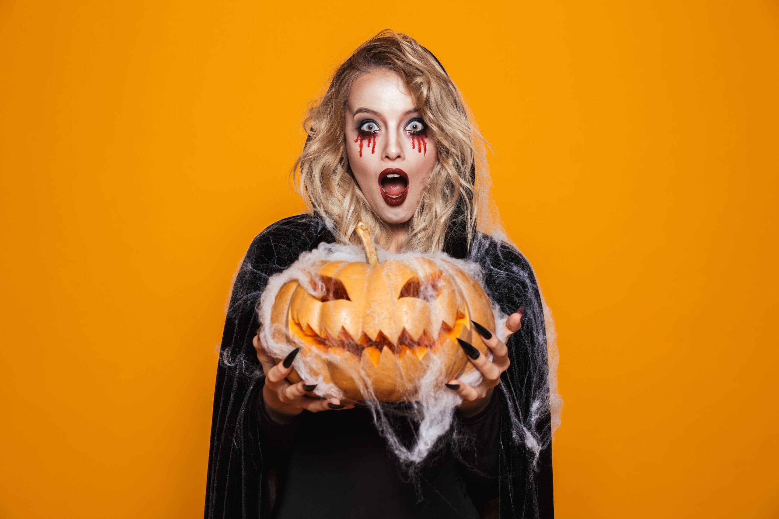 mulher vestida com traje preto e maquiagem de Halloween segurando uma abóbora esculpida, isolada em fundo amarelo.