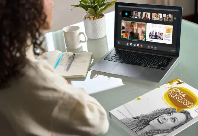 Mulher adulta, de mais ou menos 22 anos, na sua casa, assistindo uma aula online e interagindo com os colegas acompanha do material didático do curso de inglês do nível intermediário.